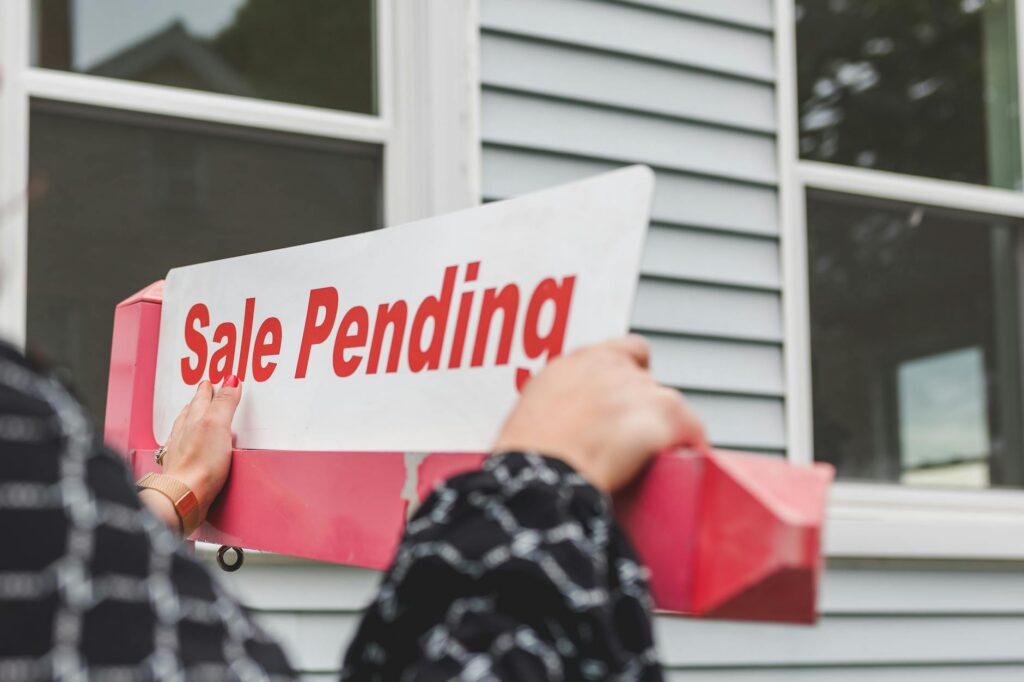 Close-up of hands holding a 'Sale Pending' sign in front of a house, featuring real estate themes.